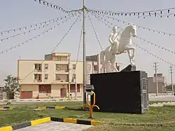 A daytime photo of a white statue of a man on horseback set upon a black blocks. Next to the statue is a pole with string lights attached in all directions. A building can be seen in the background.