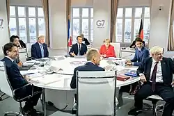 A group of seven men and one woman, sitting at a round conference table. Trump wears a dark blue suit, white dress shirt, and light blue necktie. A small sign reading "G7 France Biarritz 2019" hangs on the wall behind them.