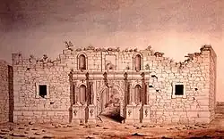 The front of a brick building; the top of the building appears torn off. Around the arched doorway are four niches, now empty. There are two small square windows.