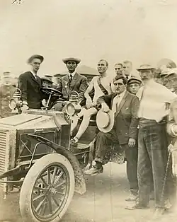 A gaunt man sits in an automobile looking exhausted, with a group of men surrounding him, looking considerably healthier. All are looking at the camera.