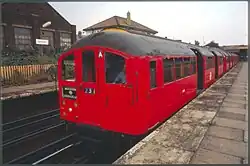 A red 1938 Bakerloo line train bound for Harrow & Wealdstone waiting at a platform at Harlesden station with its doors open
