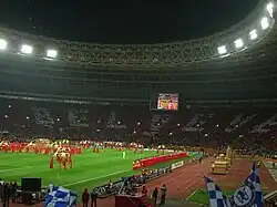 The interior of a football stadium at night, lit by floodlights. Performers are on the field and the supporters in the far stand are holding up cards that read "Believe" in silver on a red background.