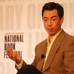 A photograph of Kyle Chayka. He is a white man with a slight widow's peak, high eyebrows and pursed lips. He is wearing an open-collared white shirt and a brow plaid suite jacket. He is in front of backdrop printed with the Library of Congress National Book Festival logo.