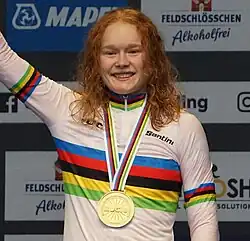 Photo of Pieterse from the waist up, with red hair just below her shoulders, with a gold medal around her neck, and wearing a rainbow jersey