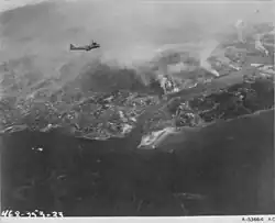 A black and white photograph which shows many clouds of smoke arising from a city which has been bombed, a plane can also be seen