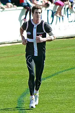 A male athlete with dark hair wearing a sleeved jersey and compression pants runs on the boundary of the grass surface of the playing arena.