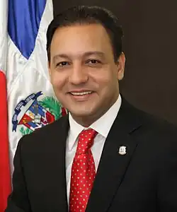 A portrait shot of a adult male smiling somewhat and looking straight ahead with Dominican Flag in the background. He has light skin and straight black hair. He wears a black suit and red tie.