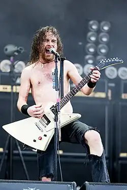 Joel, aged 34, plays guitar with both hands, while singing into a microphone on a stand. He is seen shirtless, with ripped jeans.