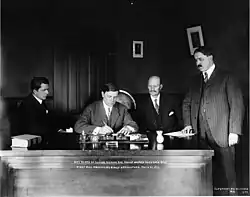 The governor sits at his desk, pen in hand, looking down at the document. The governor is flanked on the left by two men, bald one sitting, and a husky one standing. On the governor's right sits a man looking down at the document.