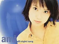 A woman standing in front of a blue shiny background, wearing a yellow t-shirt and black hair. The woman is looking towards the camera, with the woman's name and song title on the side.