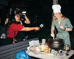 A news reporter holds her microphone to Rauwerda while she is cooking the perpetual stew while wearing a chef's hat.