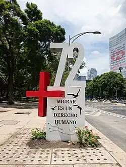 A red plus sign and the white digits "72" on a white pedestal with the Spanish words "Migrar es un derecho humano" in black uppercase letters