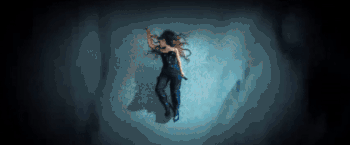 Grande lying immobile on the floor as commuters rush past her. The commuters appear blurry.