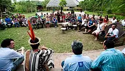 Many people are sitting on the grass, forming a circle. Most people are wearing traditional Indigenous garments
