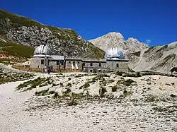 A rocky mountainous landscape has scrub brush scattered around the area. A stone building with two large silver domes is the observatory.