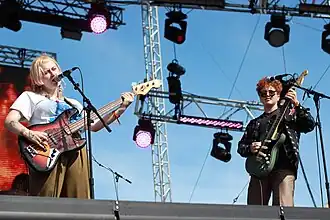 two white guitar players in their 20s perform a festival stage in the daylight — one has long blonde hair and the other has short curly red hair