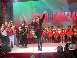 Gonzaga stands on stage wearing a red shirt, a black jacket and black pants