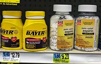 Four plastic bottles of medication on another drugstore shelf above their price tags. The two on the left are yellow with the word "Bayer" prominent in black type; above small type describes the product as "genuine aspirin". On the left are two clear plastic bottles with the Rite Aid drugstore chain logo on their yellow labels, which describe the product as "pain relief aspirin".