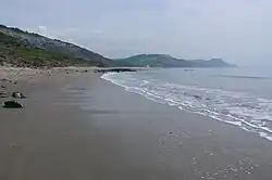 In the foreground, a vast beach of damp grey sand, lapped on the right by the waves. In the background, from the left, more or less crumbling hills slope down to the sea.
