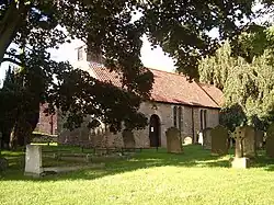 A stone church with a red tiled roof seen between trees in a churchyard. On the west gable is a bellcote.