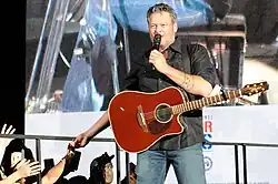 A color picture of singer Blake Shelton during a live concert, with a microphone in one hand and a red guitar strapped around him.