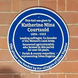 Blue plaque to commemorate Katherine Mina Courtauld 1856-1935. The plaque reads: This Hall was given by Katherine Mina Courtauld 1856-1935 Leading suffragist, Co-founder of the Women's Land Army. Renowned agriculturalist, who farmed in this parish for over 50 years. Parish & county councillor, representing Colne Engaine. This plaque was funded by the villagers of Colne Engaine.