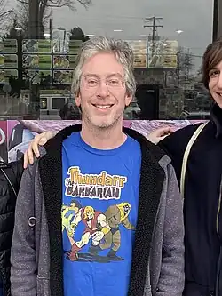 Bryan Caplan wearing a blue print t-shirt with an open, grayish hoodie over it, standing in between two people, both mostly out of the frame, grinning directly at camera