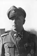 A man wearing a military uniform, peaked cap and neck order, in the shape of a cross. His cap has an emblem in shape of a human skull and crossed bones.