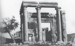A frontal black and white picture of ionic temple ruins showing a massive entrance door and a smaller side door. Two children in head covers sit at the right of the temple facade.