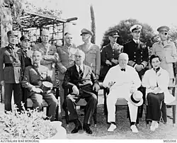A black and white grouo photograph. 3 men and a woman are seated in the front row, 8 men in different military uniforms are seen standing in the back row.