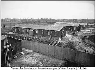 Black-and-white image of camp barracks behind a wire fence