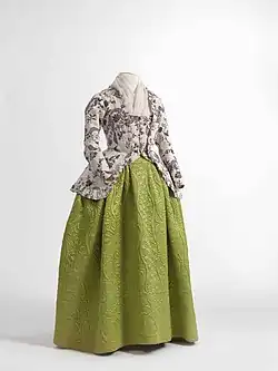 A headless mannequin is wearing a green quilted skirt and over it wearing a pink caraco with a purple fern motif. The caraco is a fitted coat that passes the hip of the wearer and buttons up the front; this version has full, fitted sleeves, a square neckline, and there is a large gap at the front skirt of the caraco.