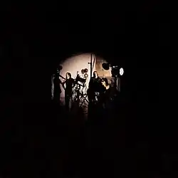 The members of Plexi in a dark room underneath a small spotlight at the center. From left to right: Barragan, Block, Angelos