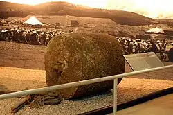 A color photograph of a stone road roller, with a plaque in front and a colorised photograph of labourers moving it in the background.