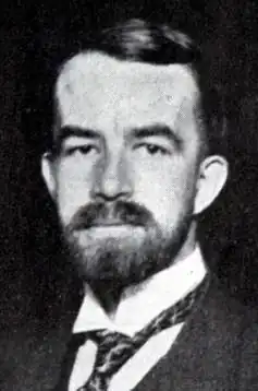 A portrait of a stern-faced bearded man in a suit and tie with a messed up collar