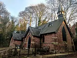 Small red brick building with three gables, arched windows and doorway, grey slate roof having a small round spire against a pale blue sky
