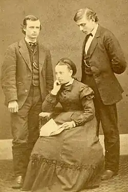 Two men standing behind a seated woman. The men are in suits and the woman is in a dress, posed with an open book on her lap.
