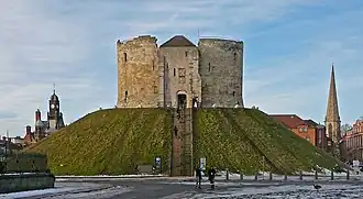 A photograph of a small castle on top of a green mound; the castle has three circular walls visible. Behind the castle the sky is overcast and dark grey.
