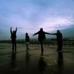 Four people standing on wet pavement against a bright sky