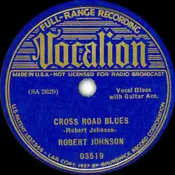 Photo of original 78 record label showing "Vocalion" in stylized lettering with "Cross Road Blues", "Vocal Blues with Guitar Acc[ompaniment]", and "Robert Johnson" as identifiers along with catalog numbers, "Made in USA", etc.