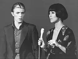 Cher, holding a microphone, wears a dark floral-patterned outfit and a short bob hairstyle. Beside her, David Bowie wears a dark suit with a buttoned-up shirt.