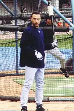 Derek Jeter stands in front of a batting cage wearing a navy long-sleeve shirt over a baseball uniform while holding a weighted bat in his left hand.