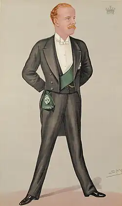 A man with blond hair and a moustache, wearing an elegant white-tie tuxedo, replete with a green sash worn beneath the jacket.