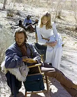 An older man in a white suit is smiling towards the left of the image. He has a black bowtie.