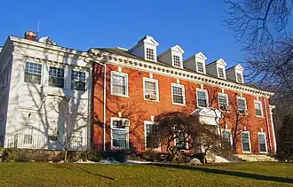 A wide brick building with dormer windows projecting from its roof and a white wooden wing on the left, seen from slightly downhill