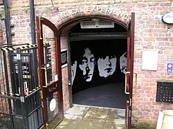 Arched doorway through a weathered red brick wall with sign "Magical Experience" above. On the wall inside the doorway is stylised silhouette of the four Beatles members