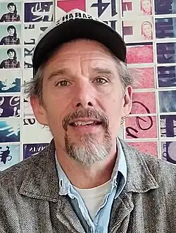 A Caucasian male aged 52 wearing a cap, with gray hair sticking out, and a graying goatie beard, wearing a gray jacket, light blue shirt and white undershirt and piercing hazel eyes