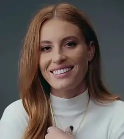 Fletcher, a White woman with a long red-brown hair, looking at the camera and smiling.