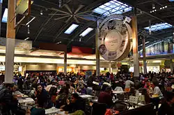 At bottom are numerous compactly-arranged tables filled with patrons eating. To the left is a large wooden support pillar, and to the right is a very large circular sign supported by a pillar at each edge; the centre of the sign contains the Vaughan Mills logo, which is surrounded on the periphery by six hand-drawn depictions of parts of the mall's interior, each separated by a small gold maple leaf. At top is a barrel-vaulted ceiling with one large skylight at its peak and two smaller skylights at its edge. Various fast food restaurants are seen in the background.