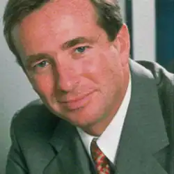 The image shows a brown-haired man in a suit. He has a slight smile and is looking into the camera.